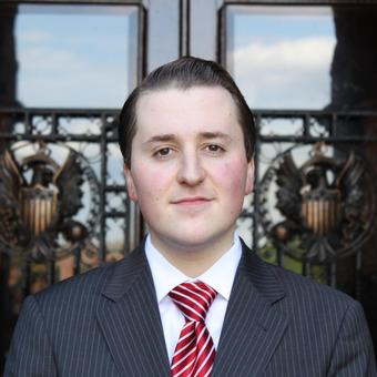 A smiling young man with brown hair wearing a gray suit, white shirt, and red tie, standing in front of Healy Hall.