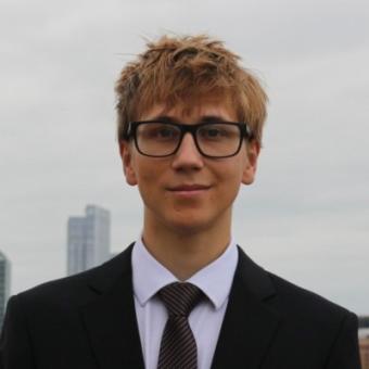 Young caucasian man with blond hair and black glasses wearing black suit, while collared shirt, and tie.