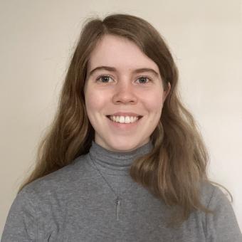 A smiling young woman with light brown wavy shoulder-length hair and brown eyes wearing a gray turtleneck standing in front of a beige wall.