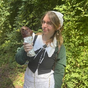 Student with glasses, white bandana, green sweater, and white collar, holding a poultry drumstick with green leaves in the background.