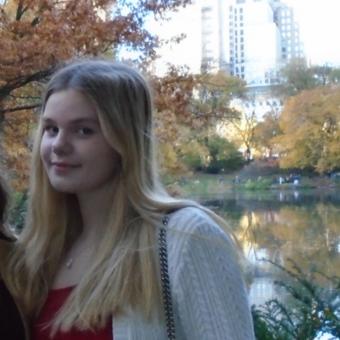 Smiling young woman with long blond hair wearing a red shirt, white sweater, and black bag, with a park in the background.
