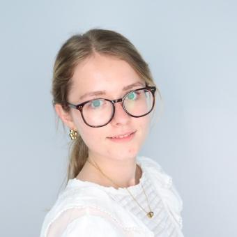 Smiling young women with blond hair and brown glasses wearing a white shirt in front of gray background.