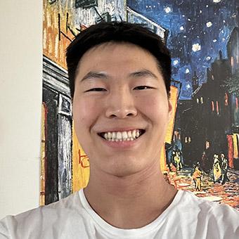 Blake Zou, a smiling student with black hair and wearing a white t-shirt, stands in front of a painting of a city