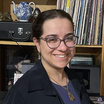 Noor Long, a smiling student wearing glasess, a blue shirt and a black blazer, stands in front of a bookshelf with LPs and other knick knackse 