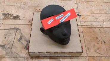 a black, 3d printed head sits on a wooden box. The head has a cavity carved out so that a triangular prism can be rotated within, showing different messages