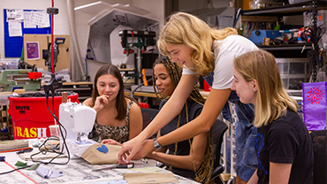 a blonde woman helping three students use a sewing machine