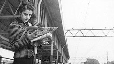 Lisa Sergio waiting on a train platform while reading a newspaper