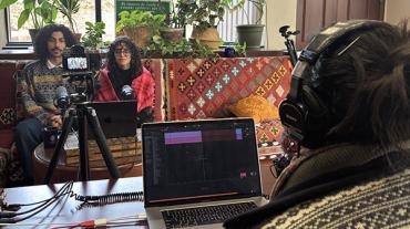 A student weraing headphones and looking at a computer screen interviews a man and a woman sitting on a sofa