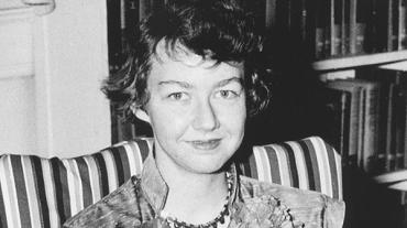 A blakc and white photo of author Flannery O'Connor, sitting on a striped chair in front of a bookshelf