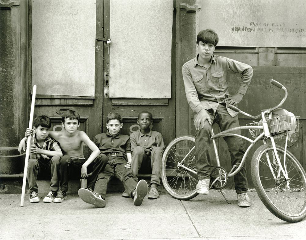 Four boys sitting in front of a door and one boy on a bicycle