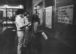 Three students stand in the lobby of Lauinger Library in 1977, reading the schedule of exams from a bulletin board
