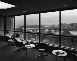 Two students seated in chairs in the 5th floor smoking lounge of Lauinger Library, with ashtrays on the tables and a sweeping view of Key Bridge and the Rosslyn skyline seen through the windows