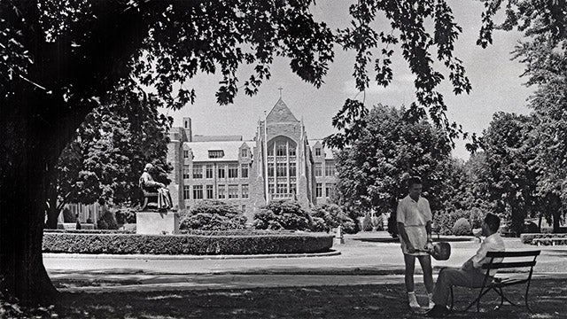 A black and white photograph from the Healy lawn looking North circa 1950. White-Gravenor Hall is in the background with the statue of John Carroll behind two men in conversation, one seated on a bench and the other standing with a tennis racquet.