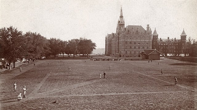 A black and white photograph from what is now Copley lawn looking south circa 1900, with Healy Hall and Old North in the background. Players in uniform are playing a baseball game, with a player at bat but no one on base.