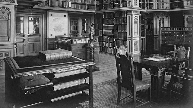 Detail from a black and white photograph of Riggs Library looking towards the main entrance, circa 1910. The two stories of the steel stacks are visible in the background, full of books, while in the foreground a stand for large atlases and dictionaries stands across from an ornate table with four similarly ornate wooden chairs.