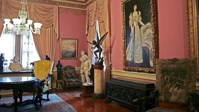 Detail from a photograph of Carrol Parlor, an art gallery formerly housed in Healy Hall, taken in 2011. The view looks towards the southeast corner of the room, where a large oil painting of Elizabeth Dhalgren hangs on the wall, and several statutes of marble and bronze rest on pedestals and the window sill. The artwork is interspersed with ornatedly carved wooden furniture, a round table and chest as well as several chairs with intricate needlework decorating the upholstery.